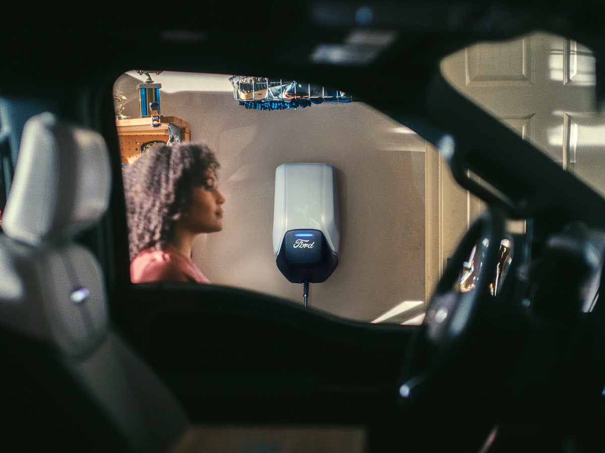 A Ford Connected Charge Station mounted on a garage wall with a woman seen through a Mustang Mach-e window.